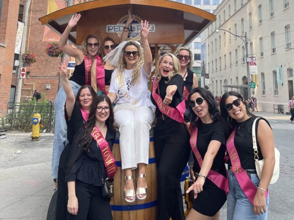 Large diverse group celebrating a private Pedal Pub tour in downtown Toronto, waving at the camera with the branded bike visible