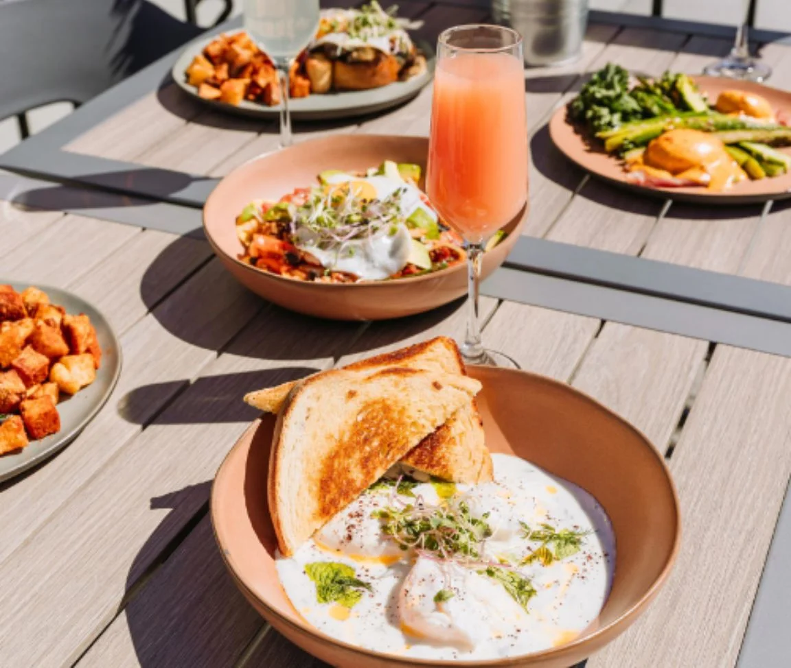 A spread of brunch dishes and mimosas laid out on an outdoor patio table at a Pedal Pub Brunch Tour stop, including poached eggs on toast, a grain bowl, Eggs Benedict, and roasted potatoes.