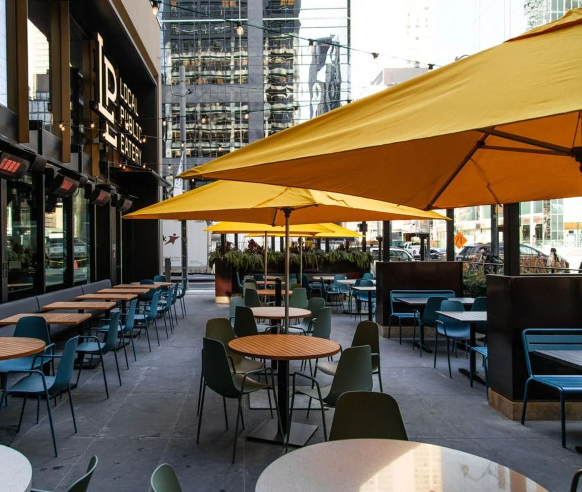 Sun-filled patio at Local Public Eatery, a Pedal Pub Brunch Tour stop in downtown Toronto, with round wood tables, teal and green chairs, and large yellow umbrellas.