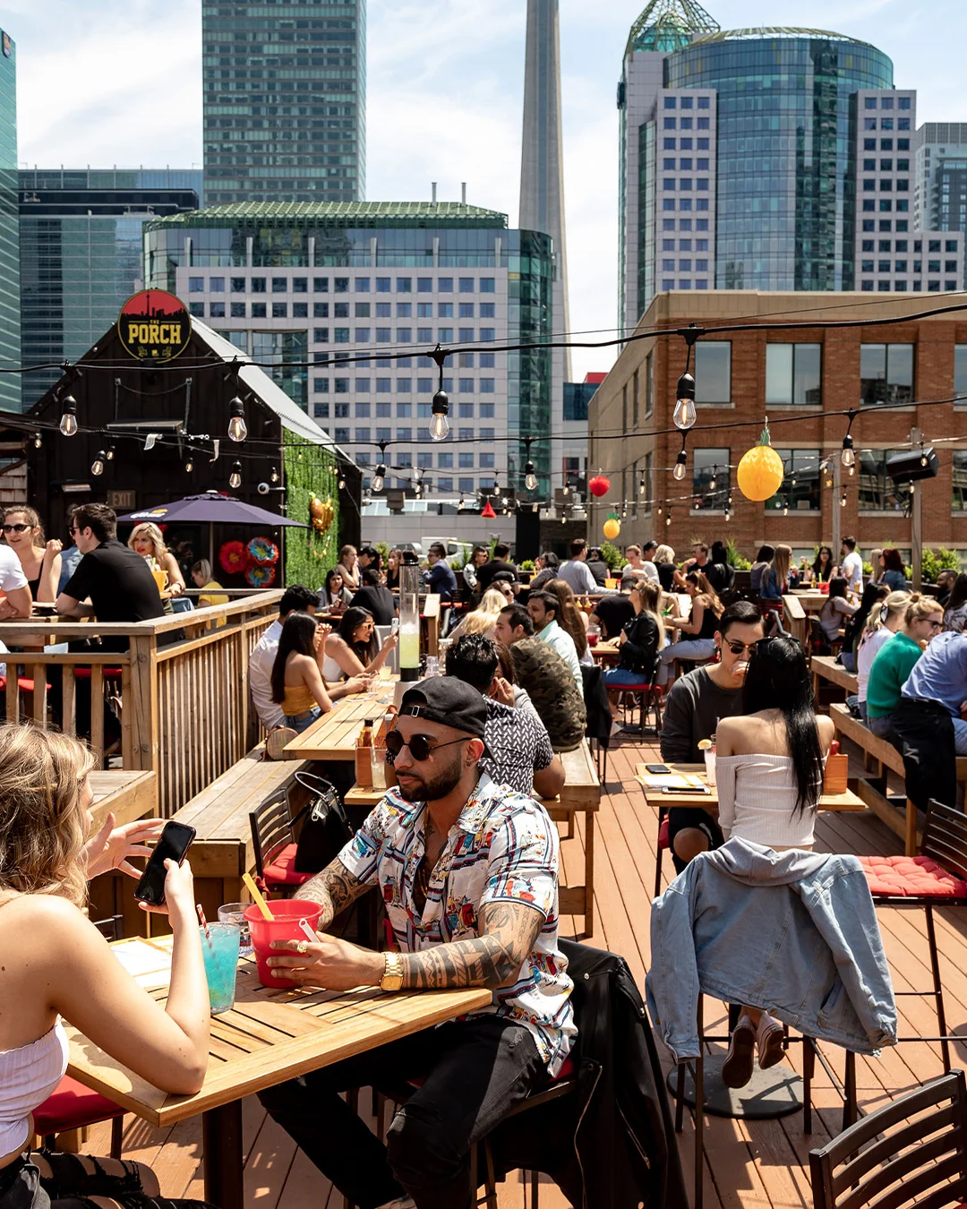 Guests enjoying drinks on the packed rooftop patio at The Porch, a Pedal Pub Brunch Tour stop in downtown Toronto, with string lights and the city skyline overhead.
