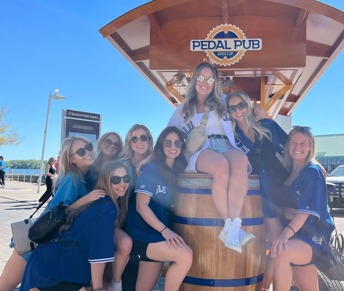 A group of women in Blue Jays gear posing around the barrel at the rear of a Pedal Pub Toronto bike at Harbourfront Centre, with Lake Ontario and a clear blue sky in the background.