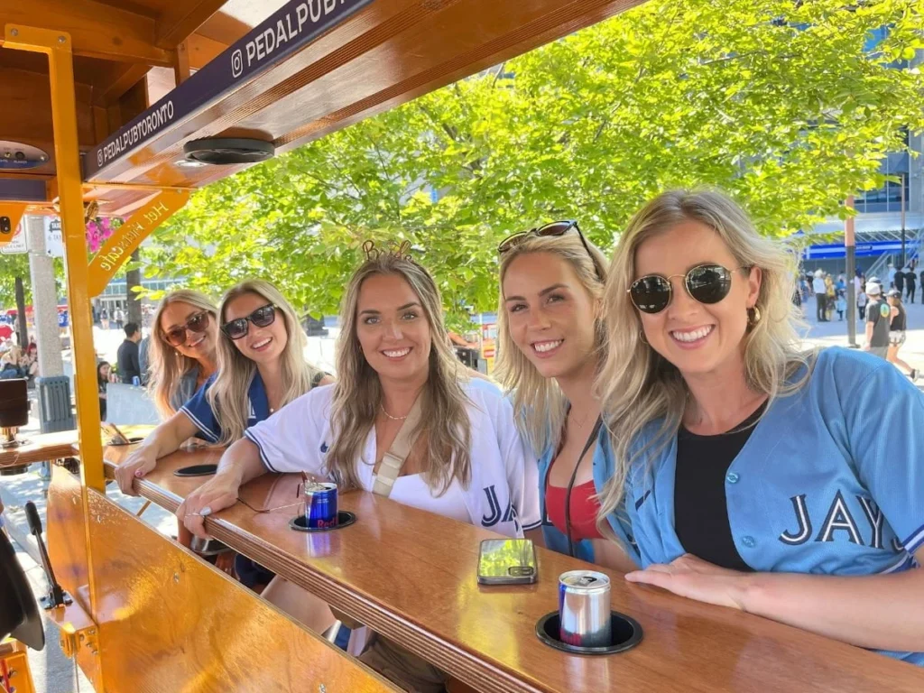 Four women in Blue Jays jerseys smile at the camera from the bar inside a Pedal Pub Toronto bike parked near Rogers Centre, with Red Bull cans in the cup holders and lush summer trees in the background.