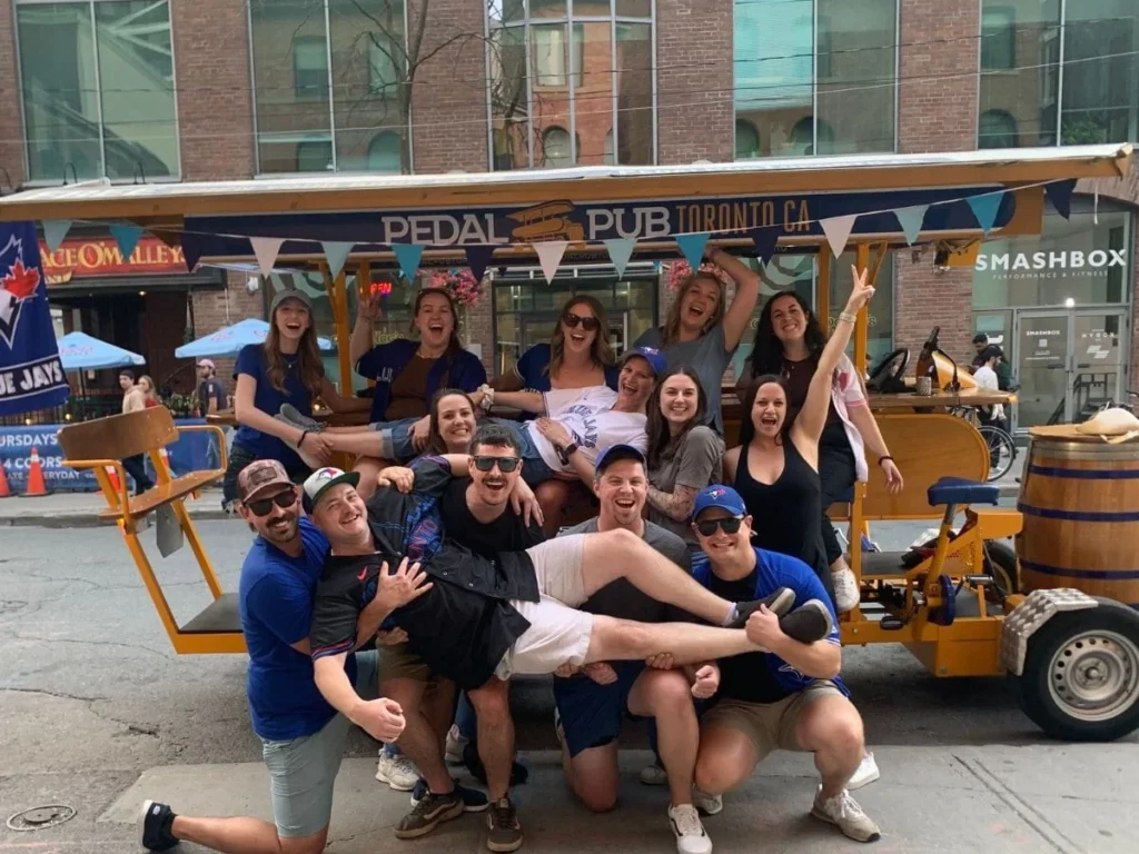A large group of Blue Jays fans erupt in celebration in front of a Pedal Pub Toronto bike on King Street West, with one person lifted in the air and a Blue Jays flag displayed on the bike.