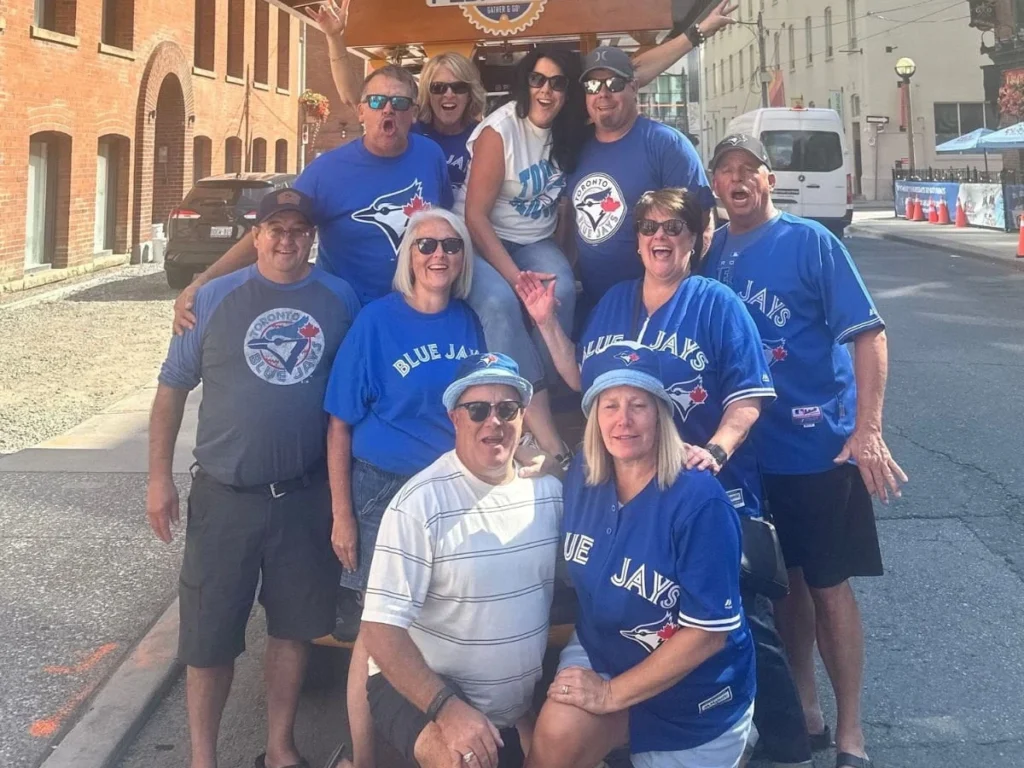 A group of enthusiastic Blue Jays fans of all ages dressed head-to-toe in Blue Jays gear posing in front of a Pedal Pub Toronto bike in the Entertainment District on a sunny afternoon.
