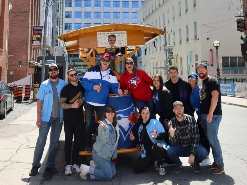 A large group of smiling friends in Blue Jays gear posing in front of a Pedal Pub Toronto bike on a downtown street with the CN Tower centered in the background on a sunny summer afternoon.