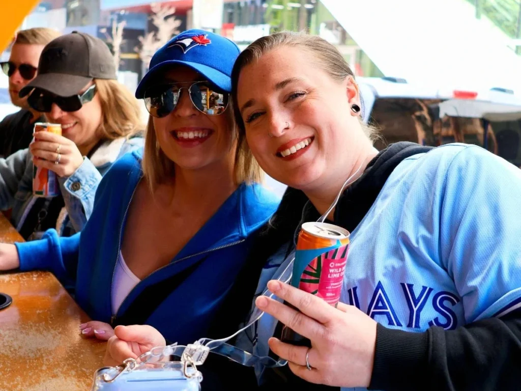 Two women in Blue Jays gear smile at the camera from inside a Pedal Pub Toronto bike, one holding a canned drink and a Fujifilm Instax camera, with blue pennant bunting overhead.