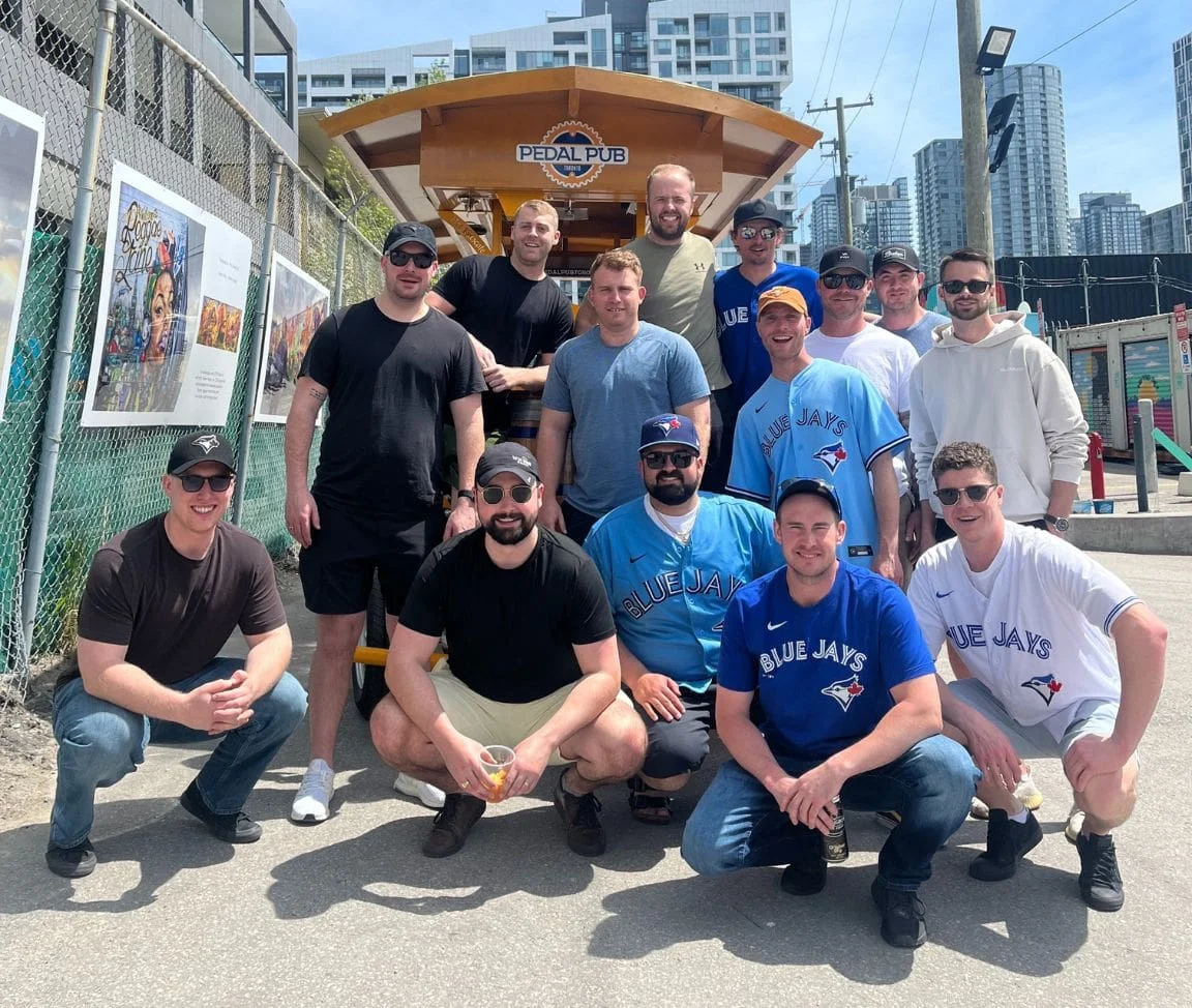 A group of men mostly dressed in Blue Jays jerseys and gear posing in front of a Pedal Pub Toronto bike near the Toronto waterfront, with the downtown condo skyline visible in the background.