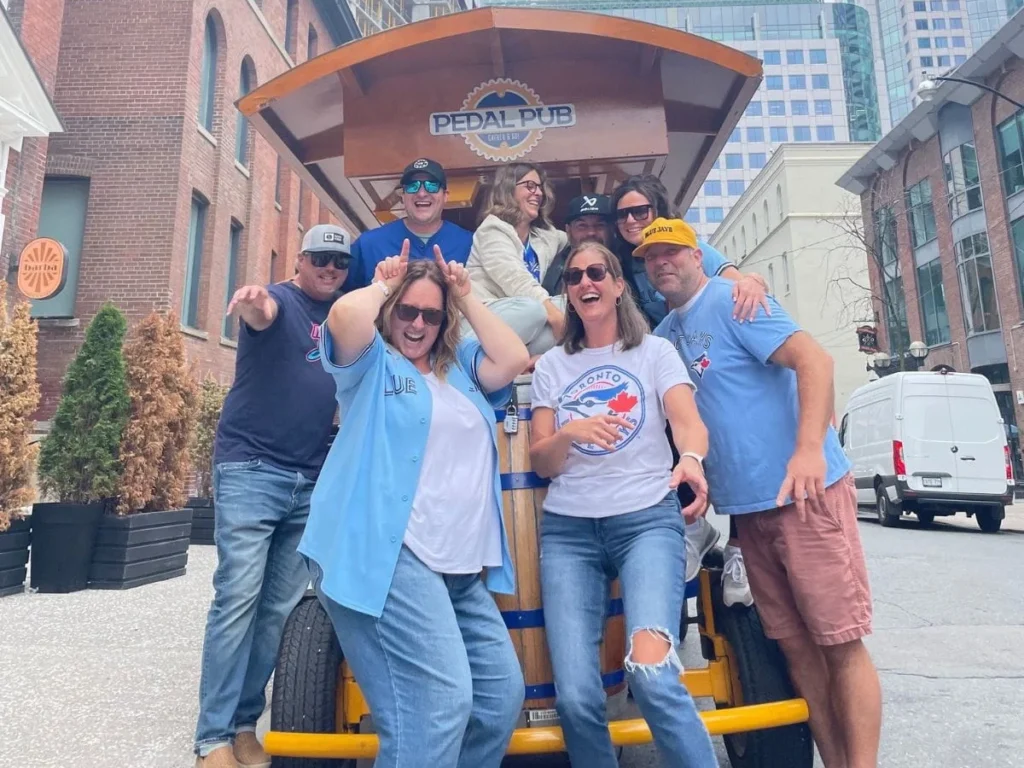A cheerful group of Blue Jays fans in jerseys and hats posing at the rear of a Pedal Pub Toronto bike in the Entertainment District, with the CN Tower and downtown skyline visible above the Pedal Pub logo on an overcast day.