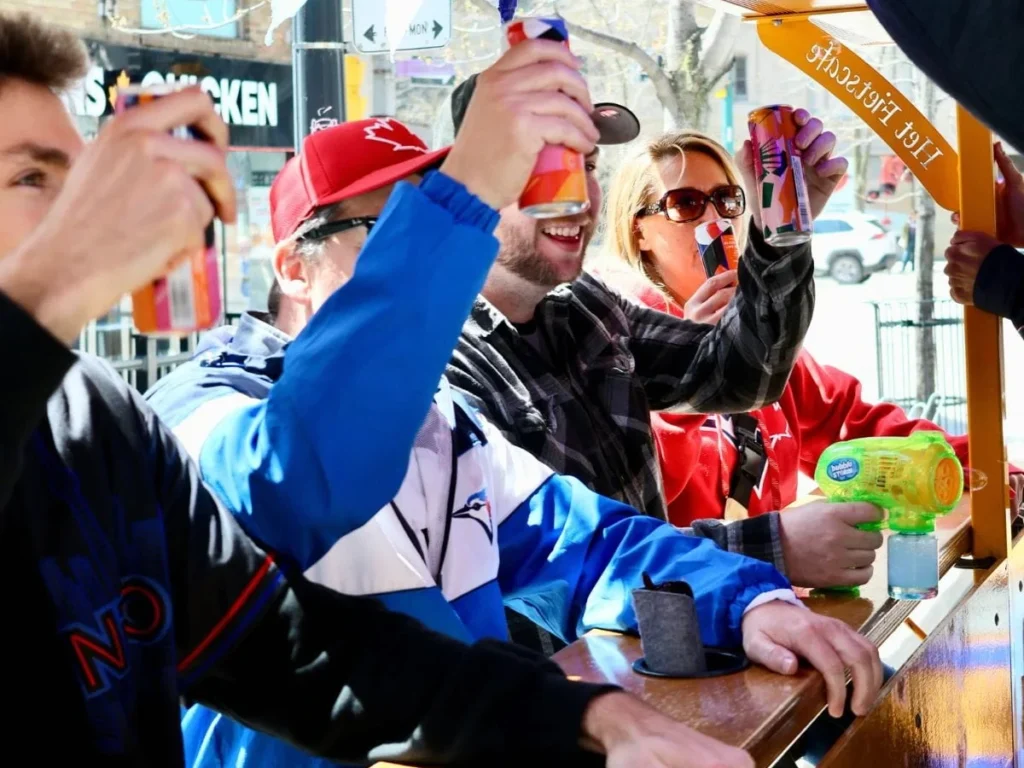 Blue Jays fans aboard a Pedal Pub Toronto bike raise their canned drinks in a toast while pedaling through downtown Toronto, with the @pedalpubtoronto handle visible on the roof rail.