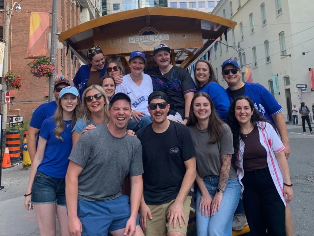 A group of friends in Blue Jays gear posing in front of a Pedal Pub Toronto bike in downtown Toronto, with the Blue Jays drum prop on the bike and the CN Tower rising above the skyline in the background.