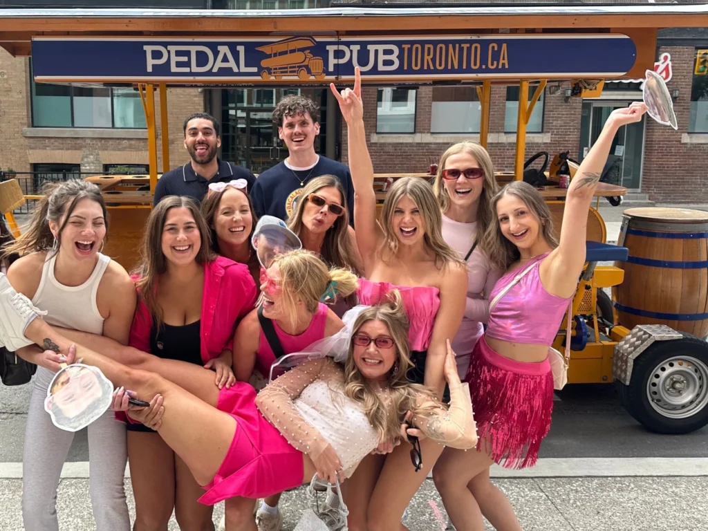 Bride in white being lifted by friends in pink outfits beside the Pedal Pub Toronto party bike