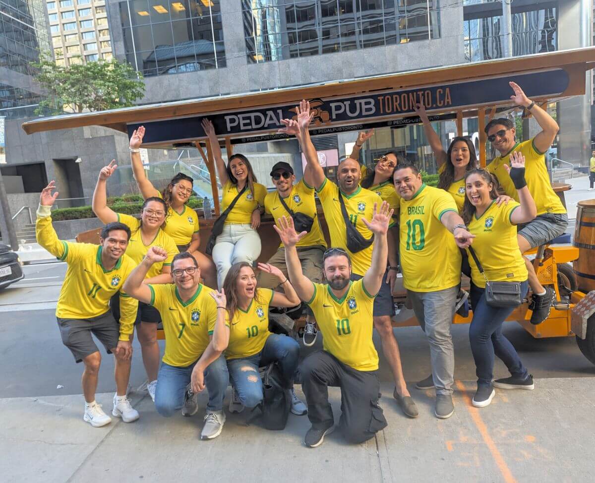 A group of Brasil supporters in matching yellow Brazil number 10 jerseys pedalling a Pedal Pub Toronto party bike through downtown Toronto, with a Brazilian flag barrel on the front of the bike.
