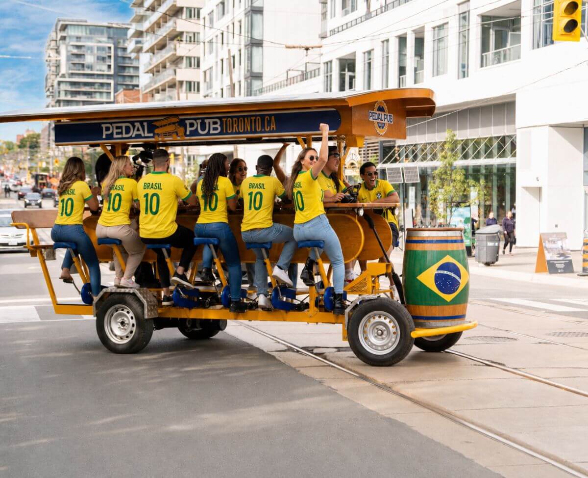 A large group of Brazil supporters in yellow Brasil jerseys celebrating in front of a Pedal Pub Toronto party bike in downtown Toronto, with arms raised and smiles all around.