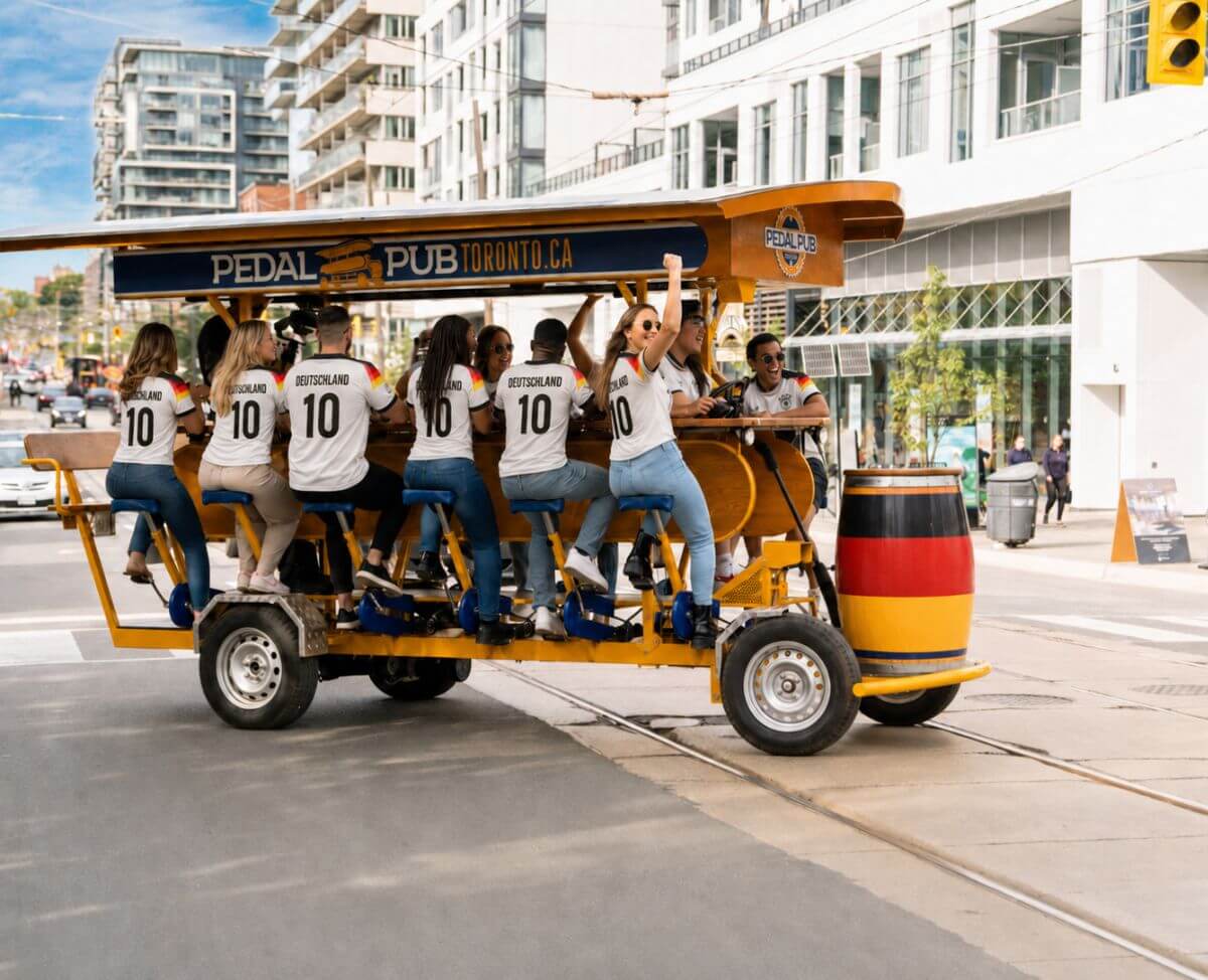 A group of Deutschland supporters in matching white Germany jerseys pedalling a Pedal Pub Toronto party bike through downtown Toronto, with a German flag barrel on the front of the bike.
