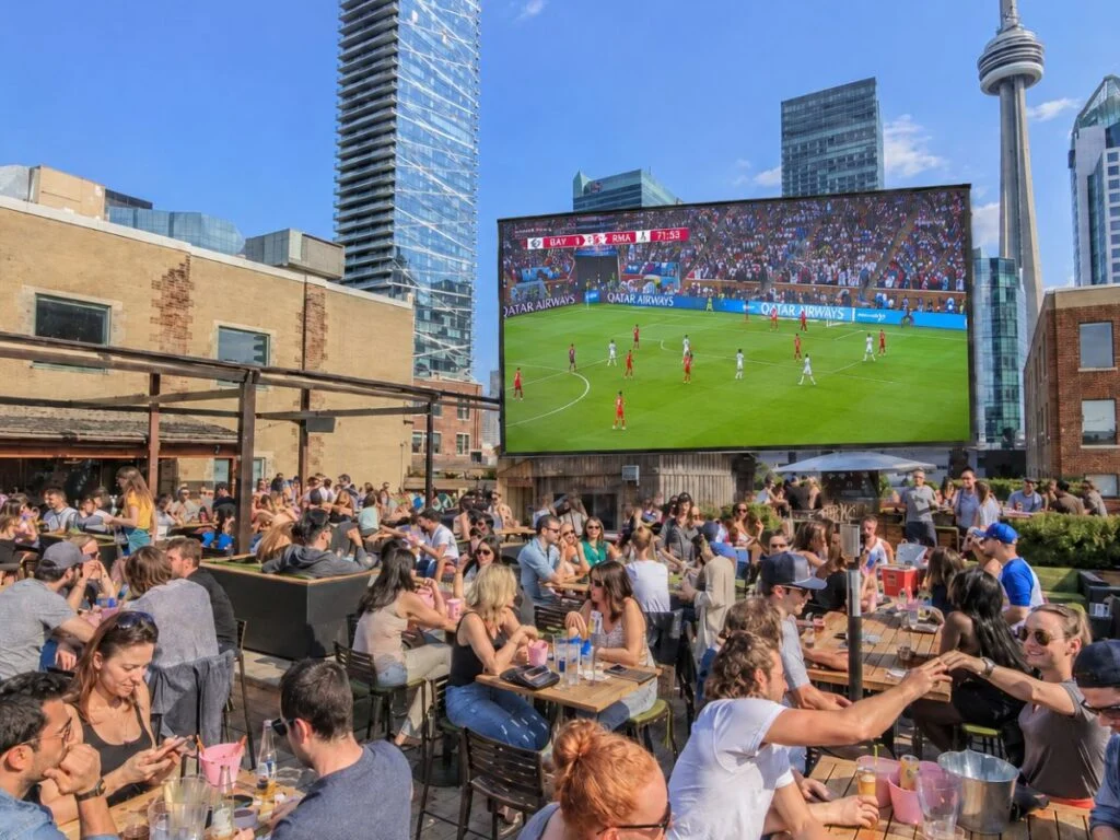 packed outdoor rooftop patio in downtown Toronto with crowds watching a football match on a massive screen, with the CN Tower and city skyline in the background on a sunny afternoon.